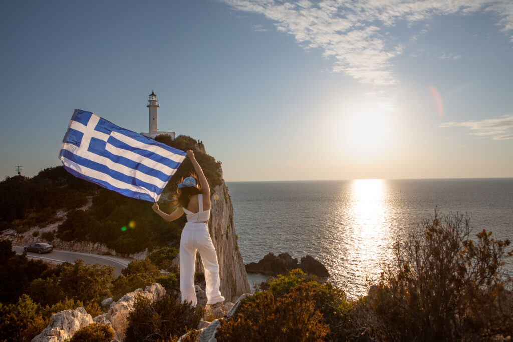 Woman holding a Greek flag stands on a cliffside over the ocean.