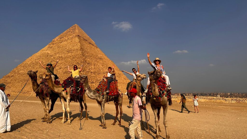 A group of women wave to the camera while riding on camels in front of pyramids on a trip to Egypt.