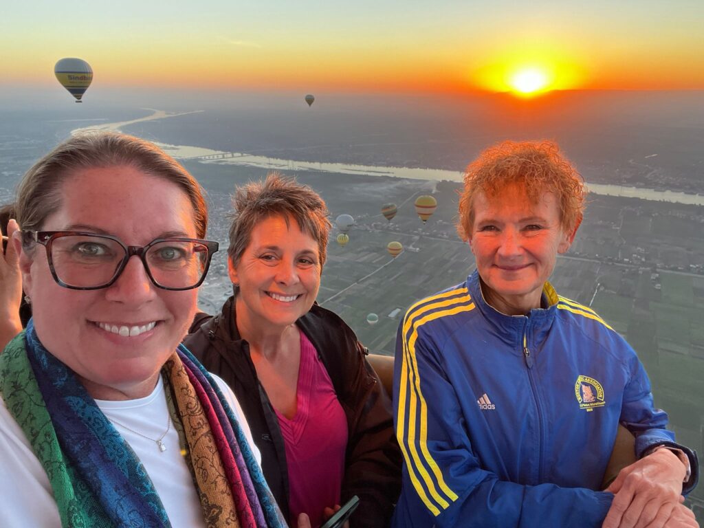 Three women are smiling during a hot air balloon ride over Luxor, Egypt with the sunrise behind them.